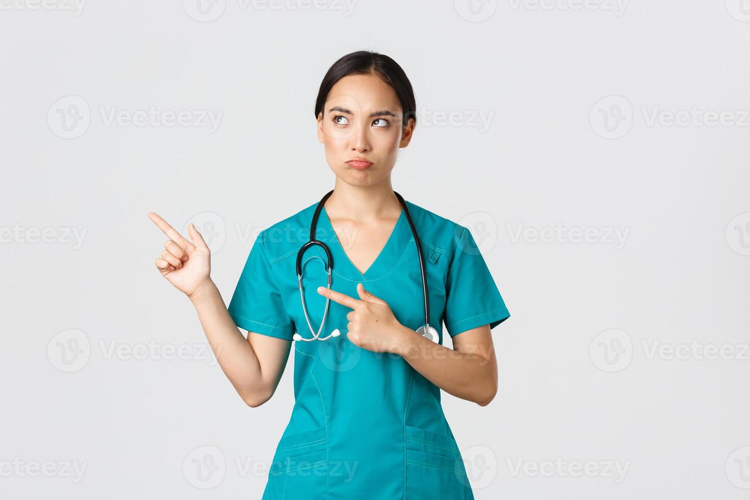 covid19, sjukvård arbetare, pandemi begrepp. besviken dyster, tjura asiatisk sjuksköterska i skrubbar, kvinna läkare ser missnöjd, pekande övre vänster hörn och rynkar dömande foto