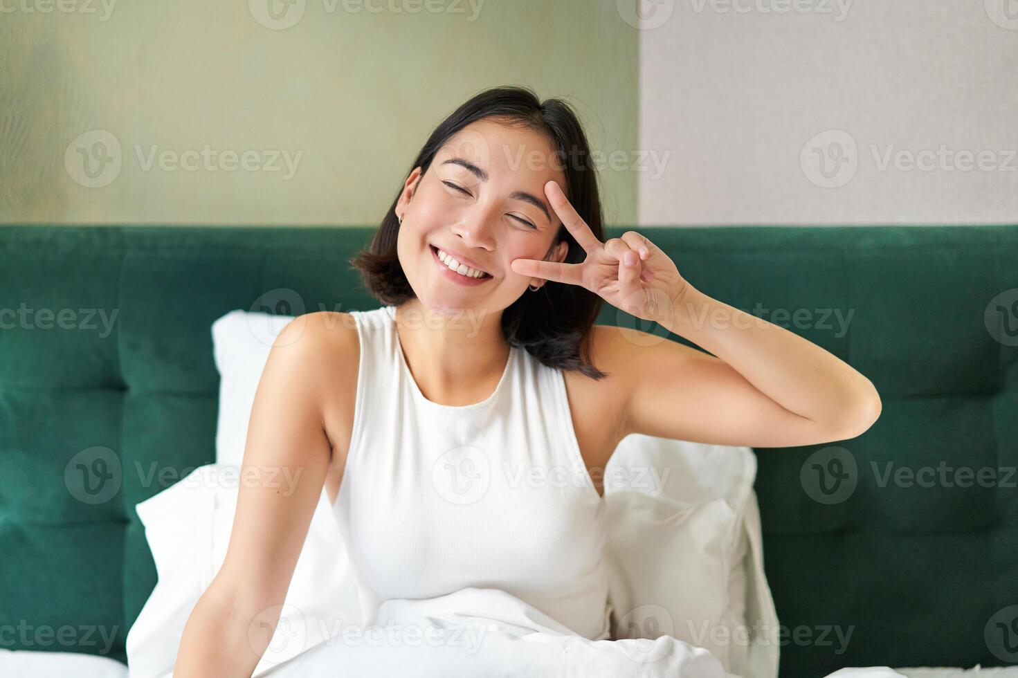 positiv asiatisk kvinna liggande i säng, som visar fred tecken, åtnjuter Lycklig morgon, vakna upp optimistisk, vistas i henne sovrum foto