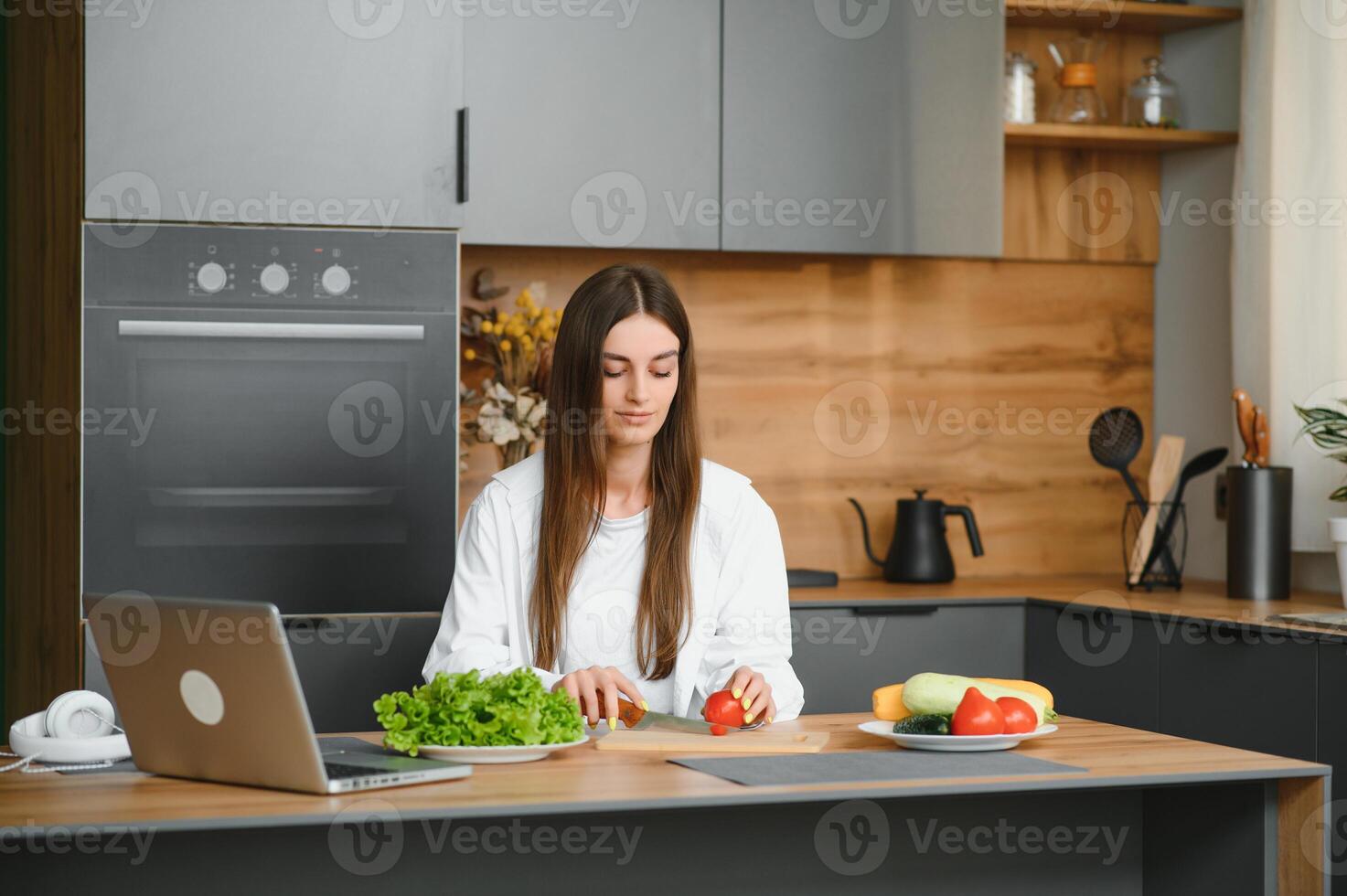 en kvinna klockor matlagning kanaler och matlagning en sallad på Hem. hon skivor grönsaker på en skärande styrelse och utseende på en bärbar dator skärm i en scandinavian stil kök. foto