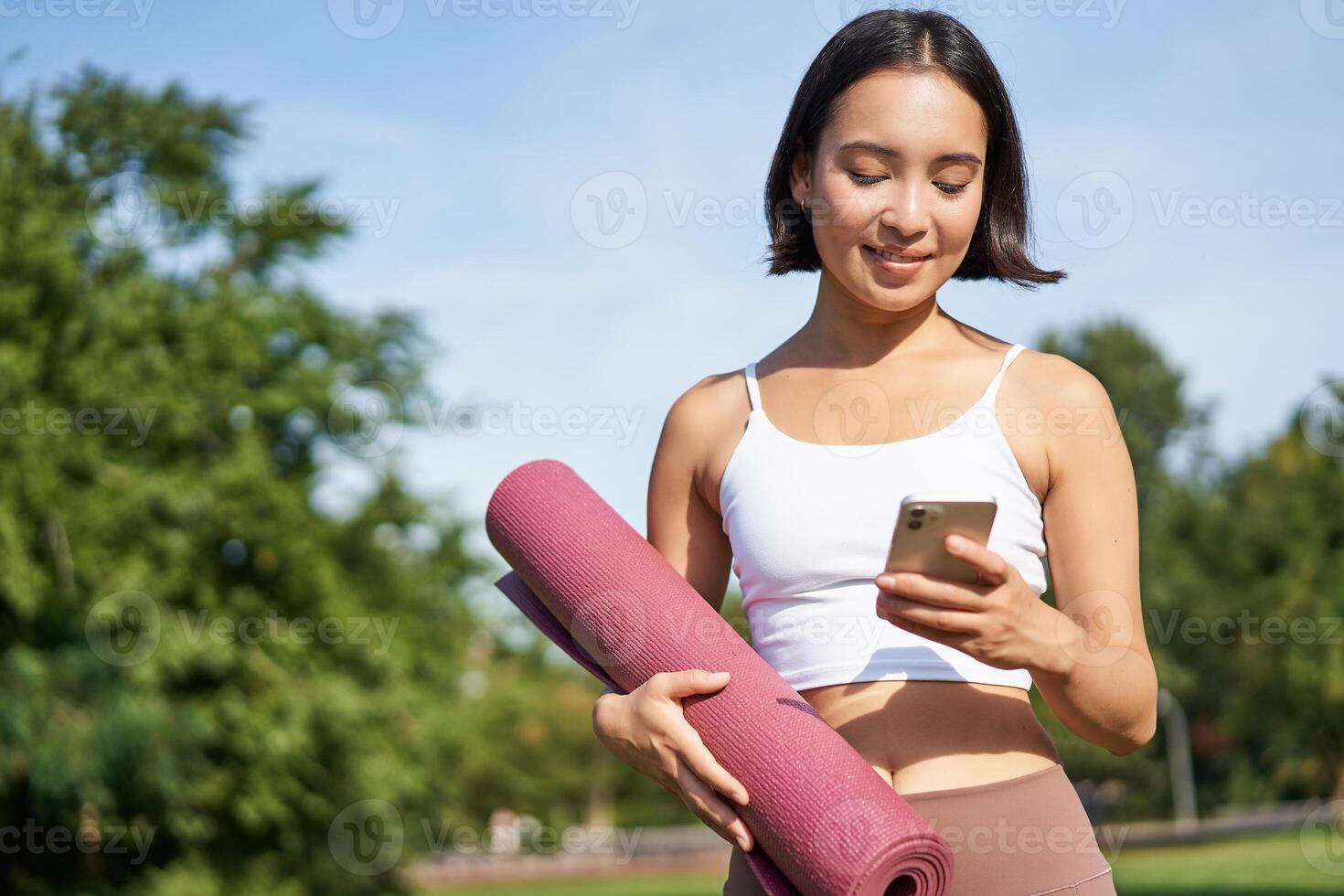 porträtt av ung asiatisk kvinna kontroll henne telefon under träna, gående i parkera med sudd matta, gående till de Gym, innehav smartphone foto