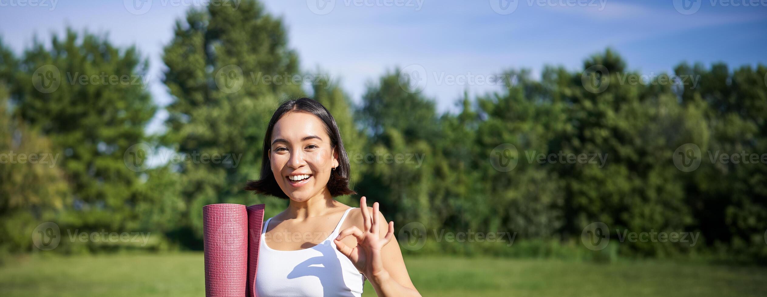 upphetsad ung kvinna stående med sporter matta, yoga kläder, visar Okej tecken, träna i parkera, välbefinnande Träning session utomhus foto