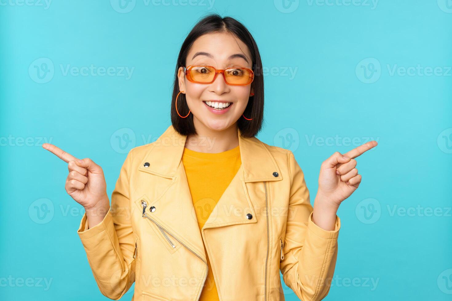 Lycklig eleganta kinesisk flicka i solglasögon, poäng fingrar åt sidan, inviterar till kolla upp ut, ser Lagra rabatt, visar vänster och höger, står över blå bakgrund foto