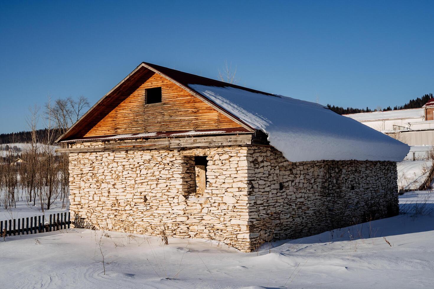 ett övergiven sten hus med en trä- staket i de mitten av en snötäckt fält på en solig dag. de tak är trä- och täckt med snö. foto