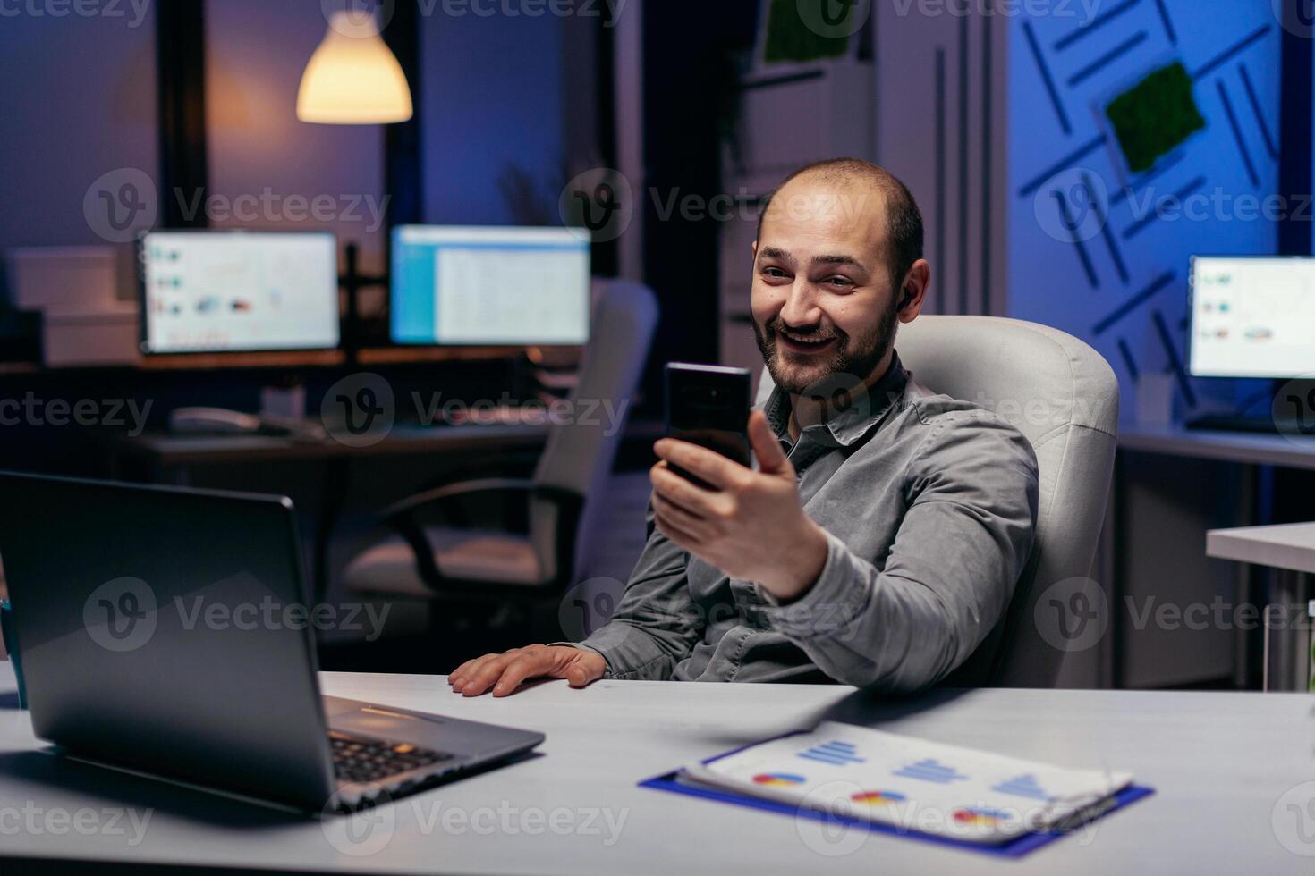 Lycklig frilansare Sammanträde på skrivbord diskuterar på uppkopplad ring upp använder sig av smartphone. affärsman i de kurs av ett Viktig video konferens medan håller på med över tid på de kontor. foto