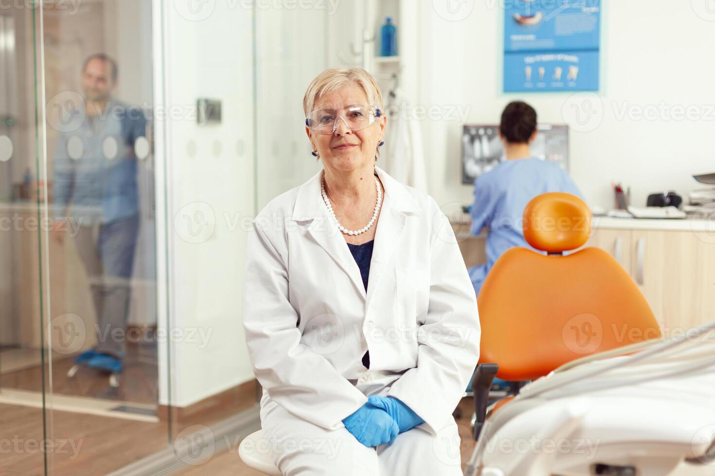 ortodontist senior i medicinsk enhetlig Sammanträde på stol ser in i kamera väntar för man pacient till Start stomatologi behandling efter tand kirurgi. team av tandläkare arbetssätt i skåp kontor foto