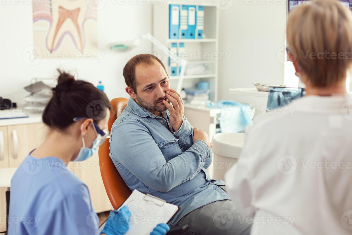 man patient med tand smärta förklara dental problem till sjuksköterska under ortodontisk utnämning. assistent skrivning anteckningar på Urklipp medan läkare tandläkare kontroll stomatologi radiografi foto