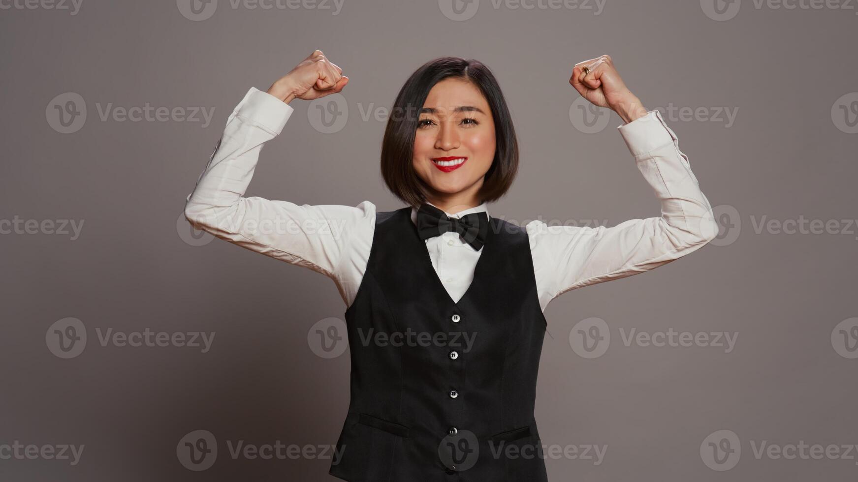 asiatisk hotell concierge böjning vapen muskler i grå studio, som visar av henne styrka och kraft i främre av kamera. receptionist med rosett och enhetlig stående med biceps böjd. kamera b. foto