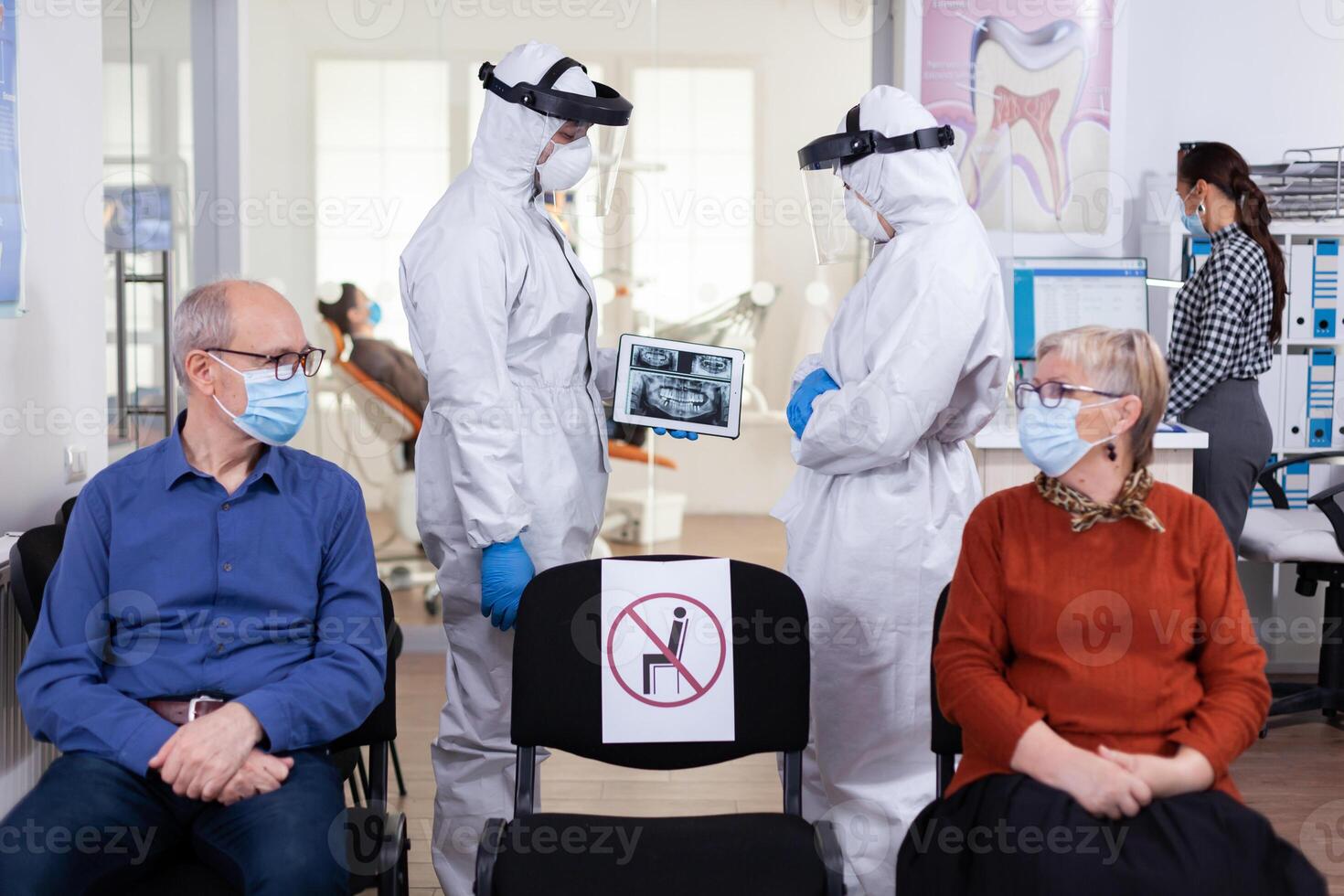 man diskuterar med sjuksköterska i dental reception bär skydd kostym mot coronavirus, äldre patienter väntar i reception förvaring distans. begrepp av ny vanligt tandläkare besök i utbrott. foto