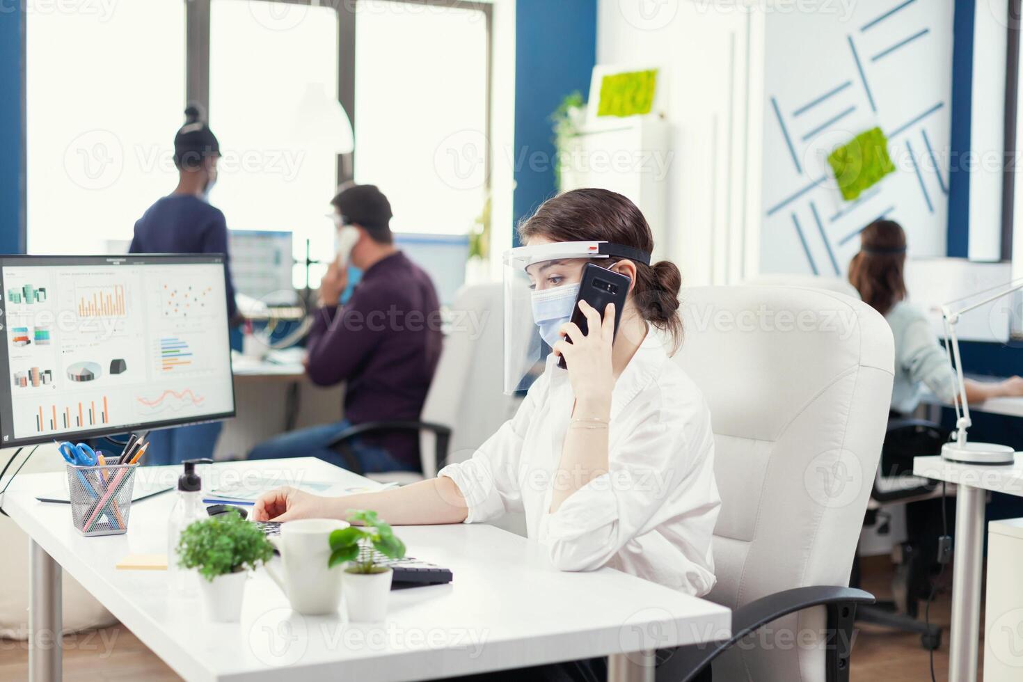 företag kvinna i kontor bär ansikte mask mot covid 19 har en konversation på smartphone. multietnisk medarbetare arbetssätt respekterar social distans i finansiell företag. foto