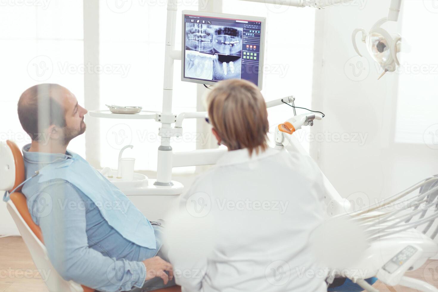 senior stomatolog kvinna diskuterar med sjuk man handla om sjukvård behandling ser på tand behandling under stomatologi sjukhus kontor. ortodontisk läkare granskning dental röntgen foto