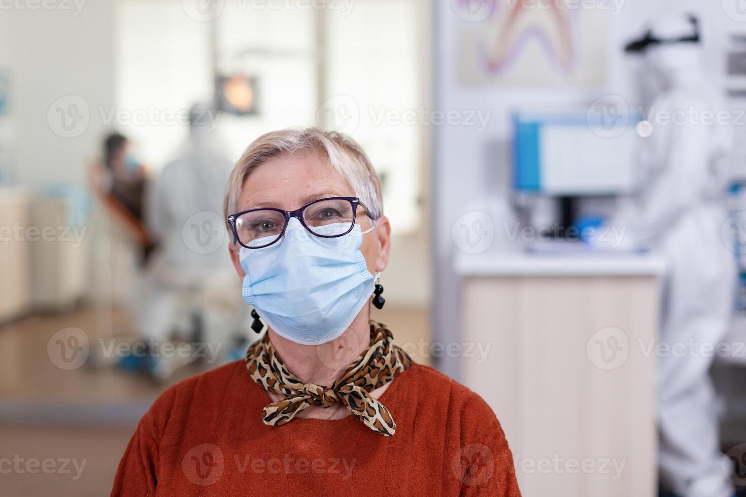 porträtt av pensionerad patient i dental kontor ser på kamera bär ansikte mask Sammanträde på stol i väntar rum klinik medan läkare arbetssätt. begrepp av ny vanligt tandläkare besök i coronavirus utbrott. foto