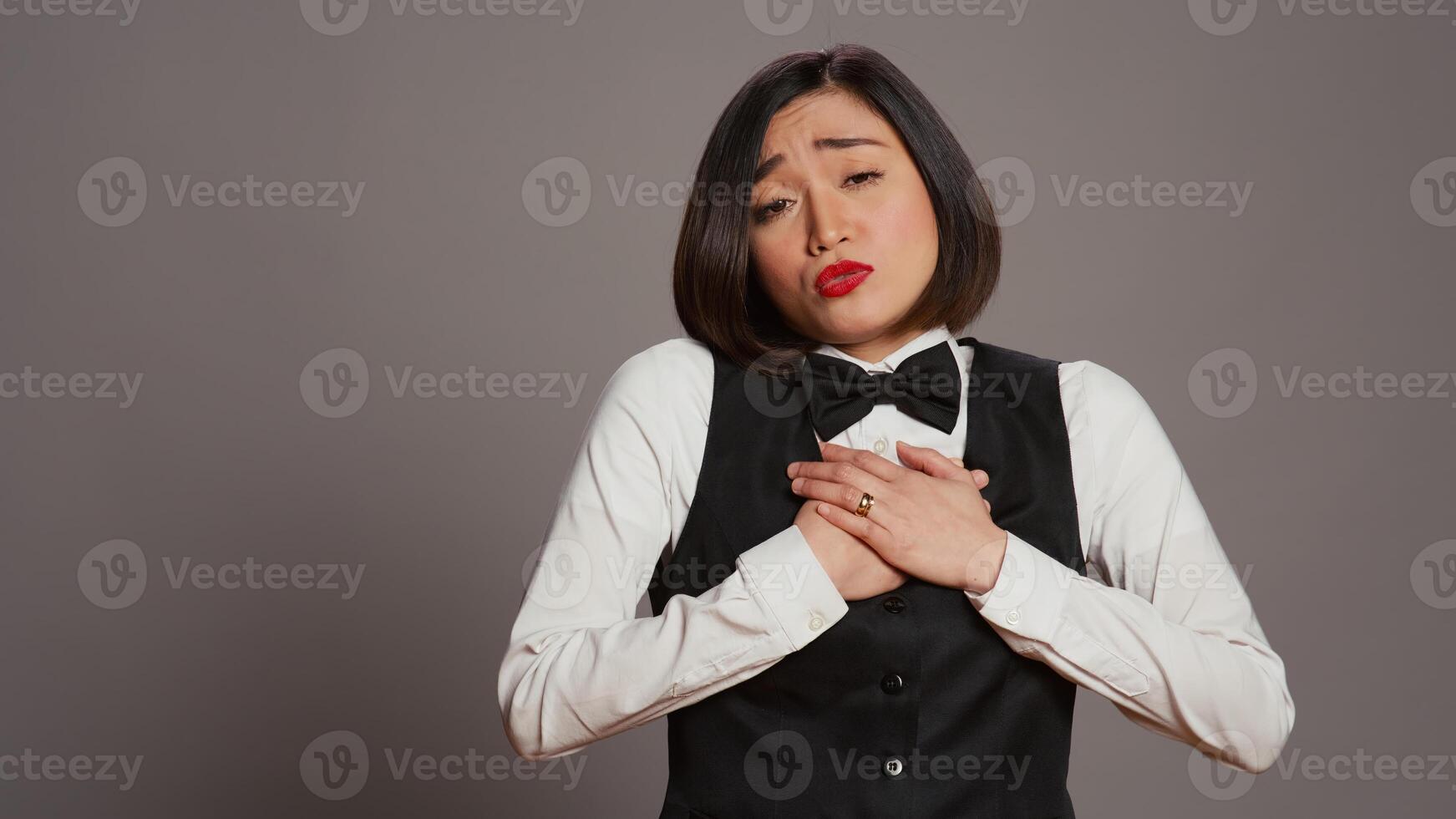 främre skrivbord personal känsla uttråkad och sömnig på kamera, som handlar om med burnout och förvirring över grå bakgrund. receptionist med enhetlig och slips varelse utmattad och osäker. kamera b. foto