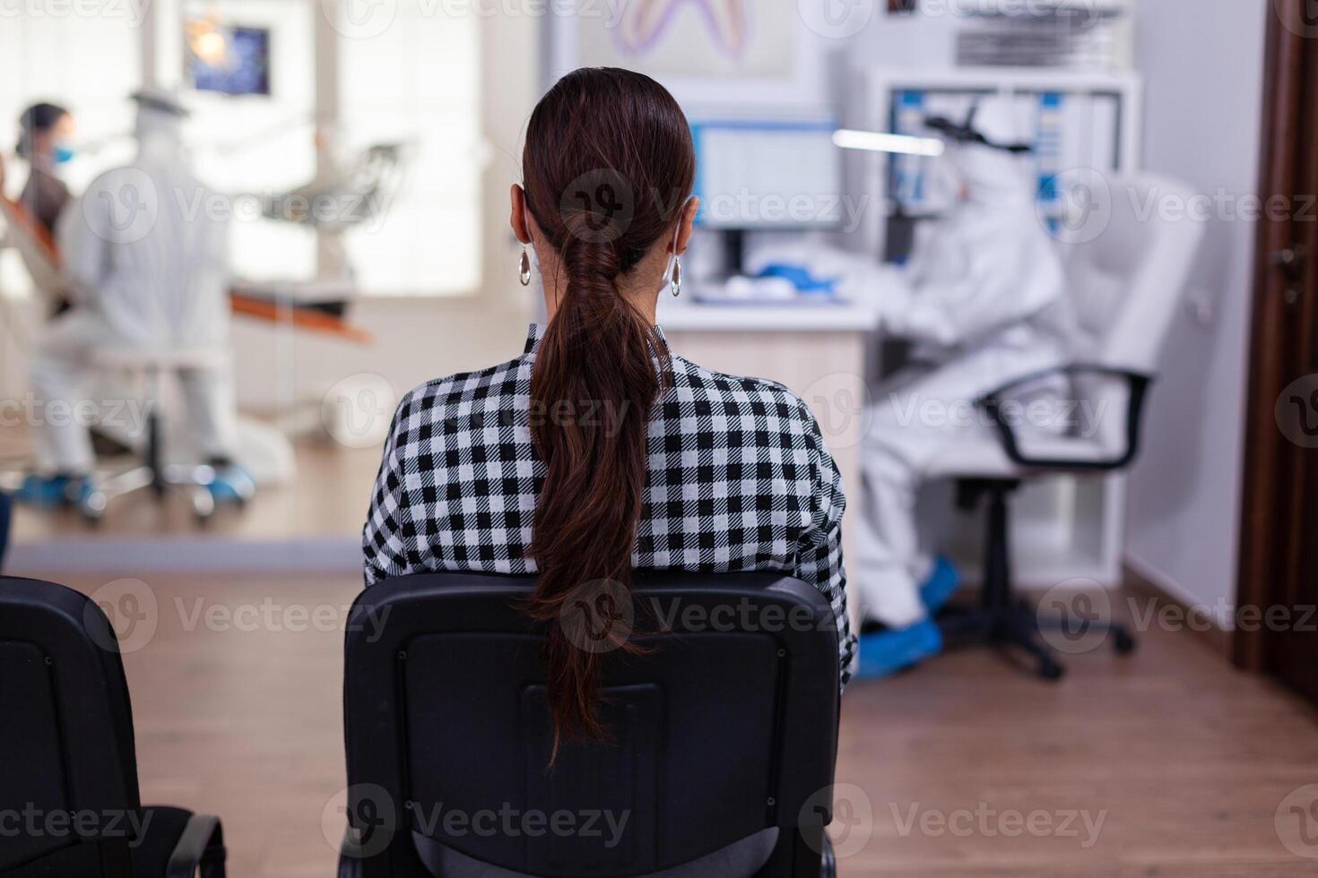 tillbaka se av kvinna i tandläkare kontor väntar för samråd Sammanträde i stolar på väntar rum, assistent i ppe kostym fyllning papper arbete på dator, tandvård läkare hört patient. foto