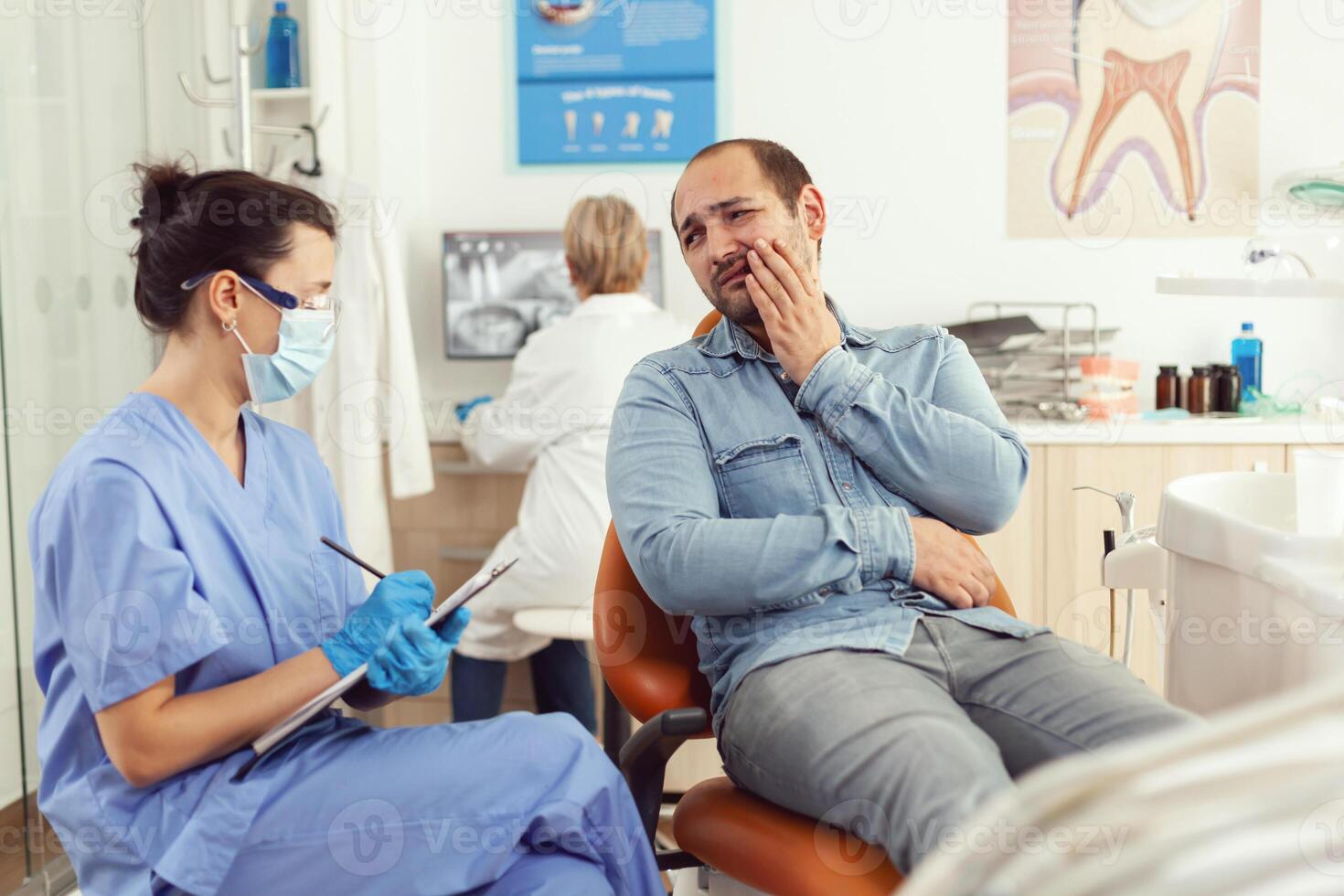 stomatolog sjuksköterska förklara medicinsk procedur till sjuk patient medan Sammanträde i stomatologisk kontor rum. assistent tar anteckningar på Urklipp talande med man framställning för dental samråd foto