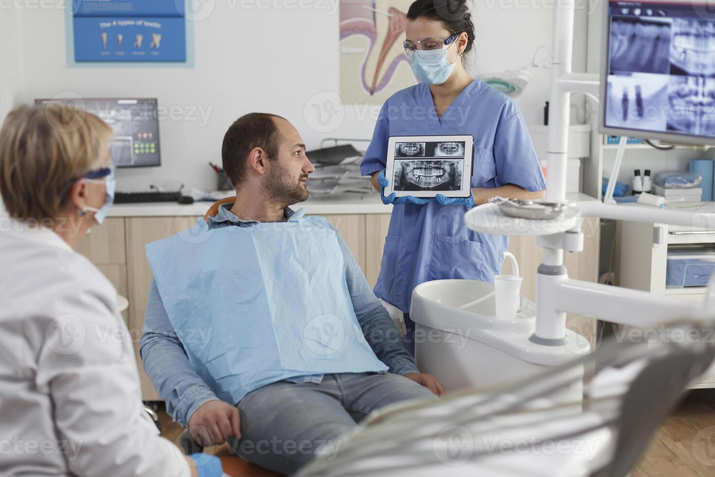 tandläkare assistent med ansikte mask innehav Tabet dator förklara tänder radiografi till sjuk patient under tandvård samråd i stomatologisk kontor rum. team arbetssätt på karies behandling foto