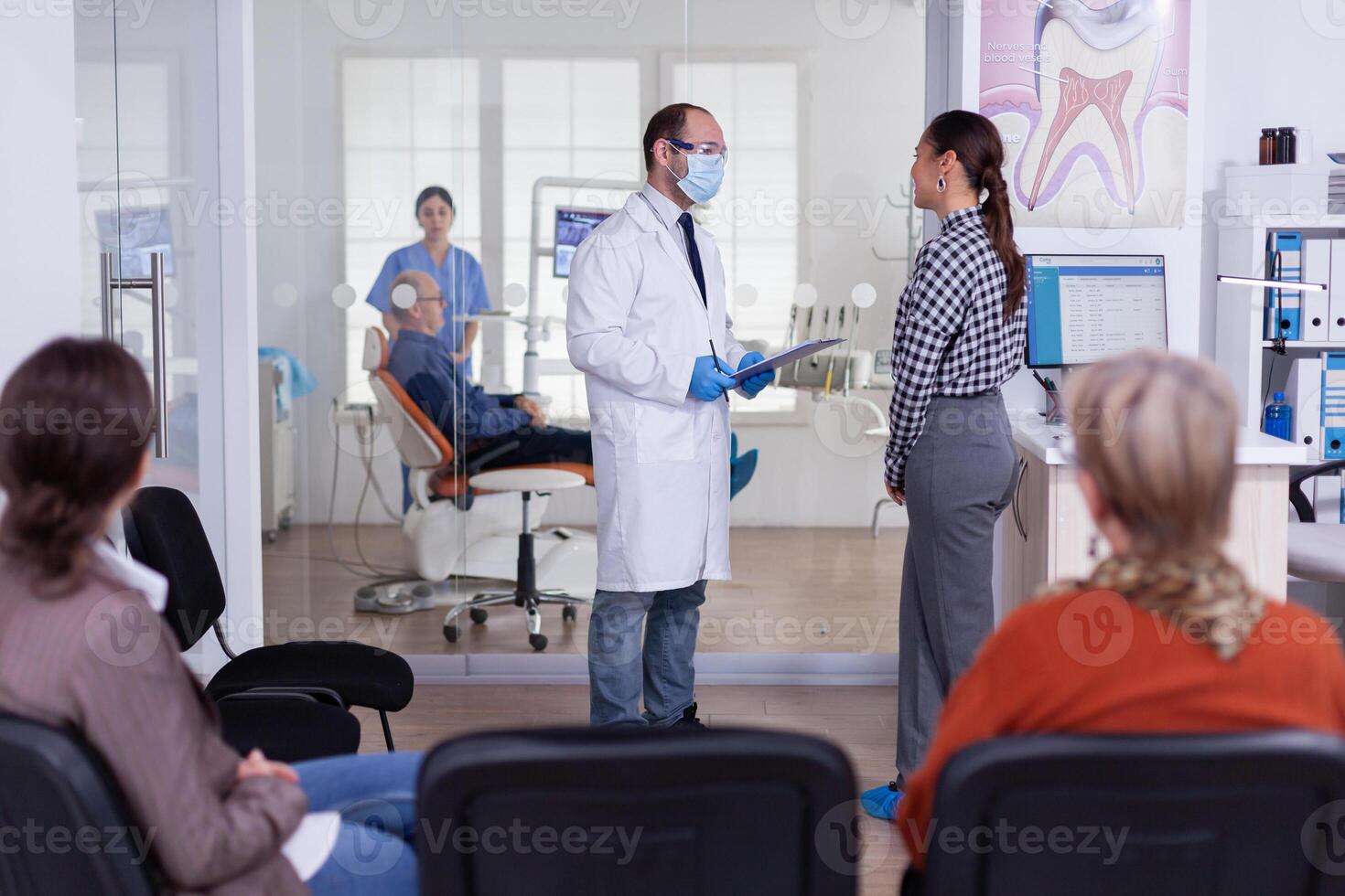 tandläkare läkare förhör kvinna och tar anteckningar på Urklipp stående i väntar område. ung patient förklara dental problem till stomatolog tala i fullt med folk reception av klinik. foto