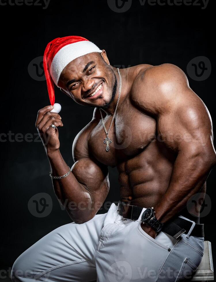 afrikansk amerikan man i jul hatt. attraktiv man med naken magmuskler Sammanträde. santa claus hatt. foto