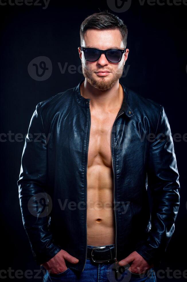 ung stilig macho man i solglasögon med öppen läder jacka avslöjande muskulös bröst och torso, ser till de kamera. närbild foto