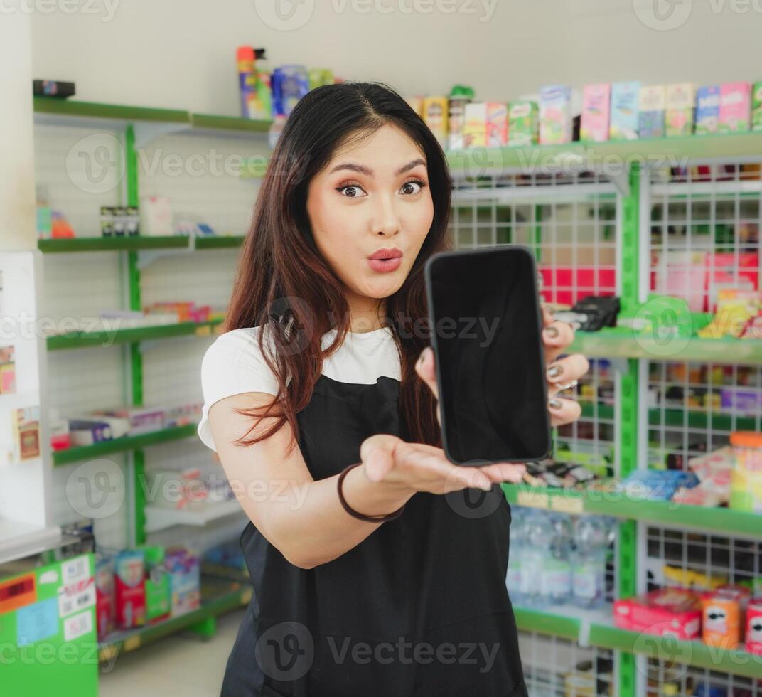 upprörande asiatisk kvinna som en kassör är bär svart förkläde och som visar henne telefon skärm foto