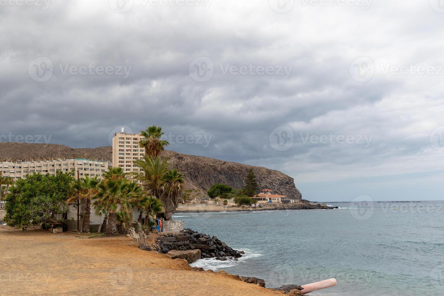 kust landskap med strand, handflatan träd, och hotell mot en bakgrund av klippor under en molnig himmel i los cristianos, teneriffa. foto