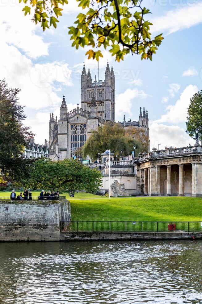 historisk katedral med spiror Bakom träd, tittade tvärs över en flod med människor avkopplande förbi de vatten på en solig dag i bad, england. foto