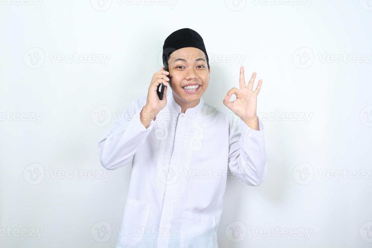 leende muslim asiatisk man visa Okej tecken medan talande på hans telefon med leende ansikte isolerat foto