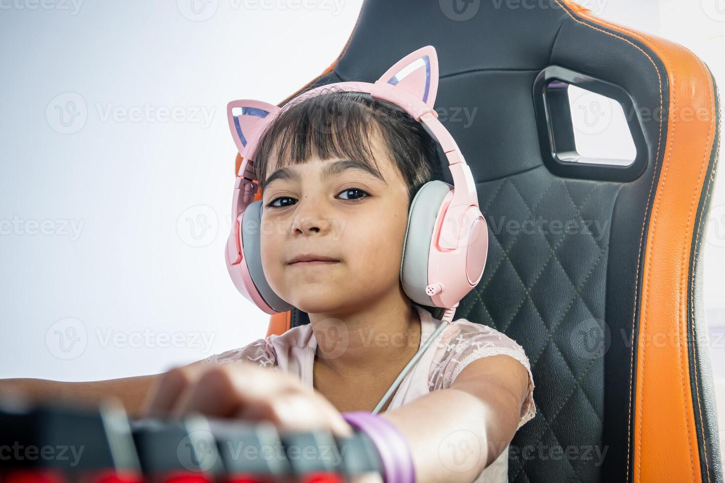 porträtt av en liten flicka med pc gamer Tillbehör. hon utseende på de kamera. hon bär hörlurar med katt öron och är Sammanträde i en gaming stol i främre av de tangentbord. foto