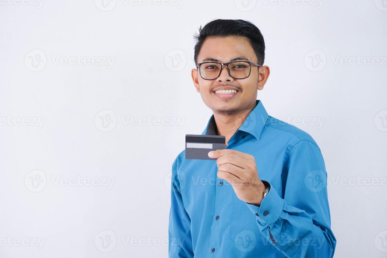 leende eller Lycklig ung asiatisk affärsman med glasögon innehav kreditera kort bär blå skjorta isolerat på vit bakgrund foto