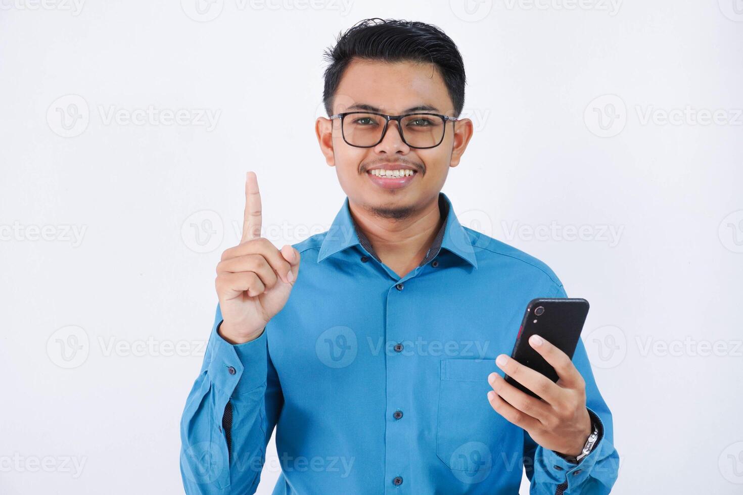 leende anställd asiatisk man med glasögon innehav telefon med finger pekande upp bär blå skjorta isolerat vit bakgrund foto
