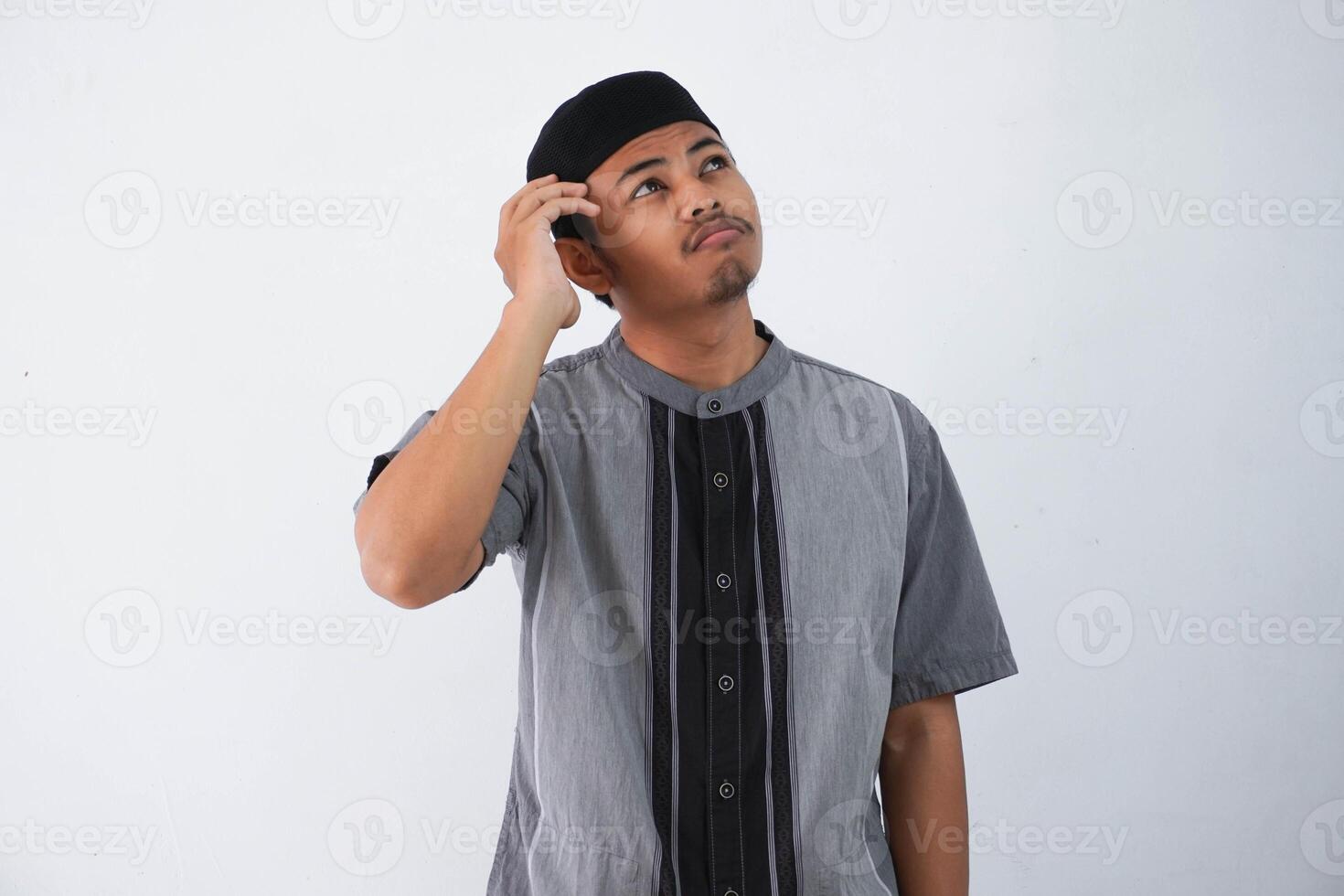 tänkande ung asiatisk muslim man innehav huvud tänkande gest, ser för aning bär grå muslim kläder isolerat på vit bakgrund foto