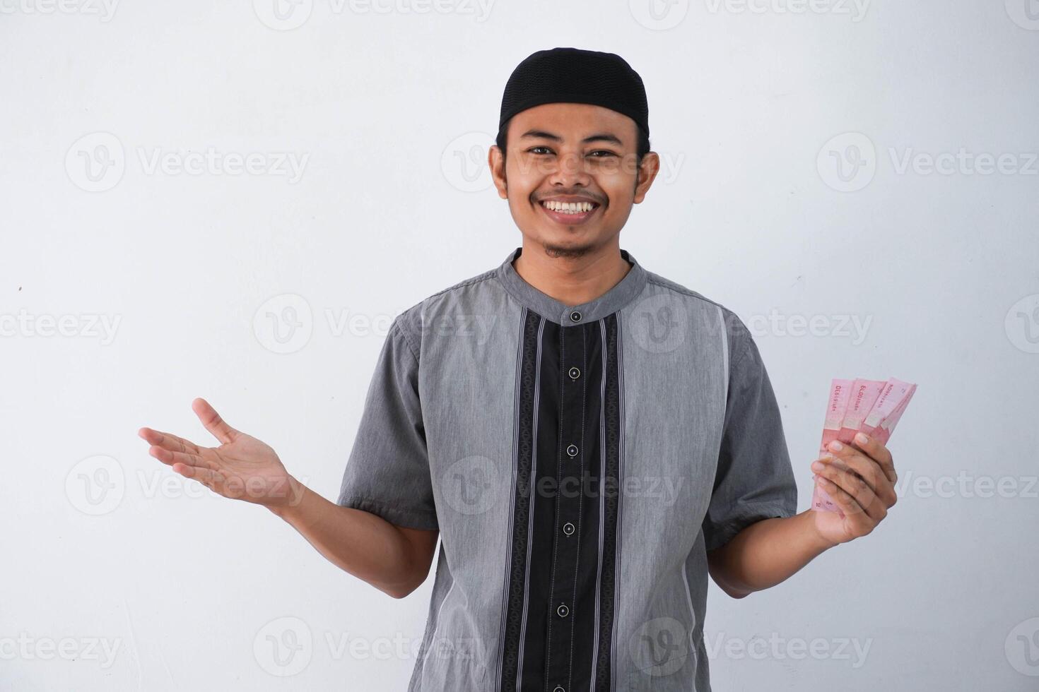 leende eller Lycklig ung asiatisk muslim man innehav och pekande papper pengar gåva thr från familj ramadan eid al-fitr tid bär grå muslim kläder isolerat på vit bakgrund foto