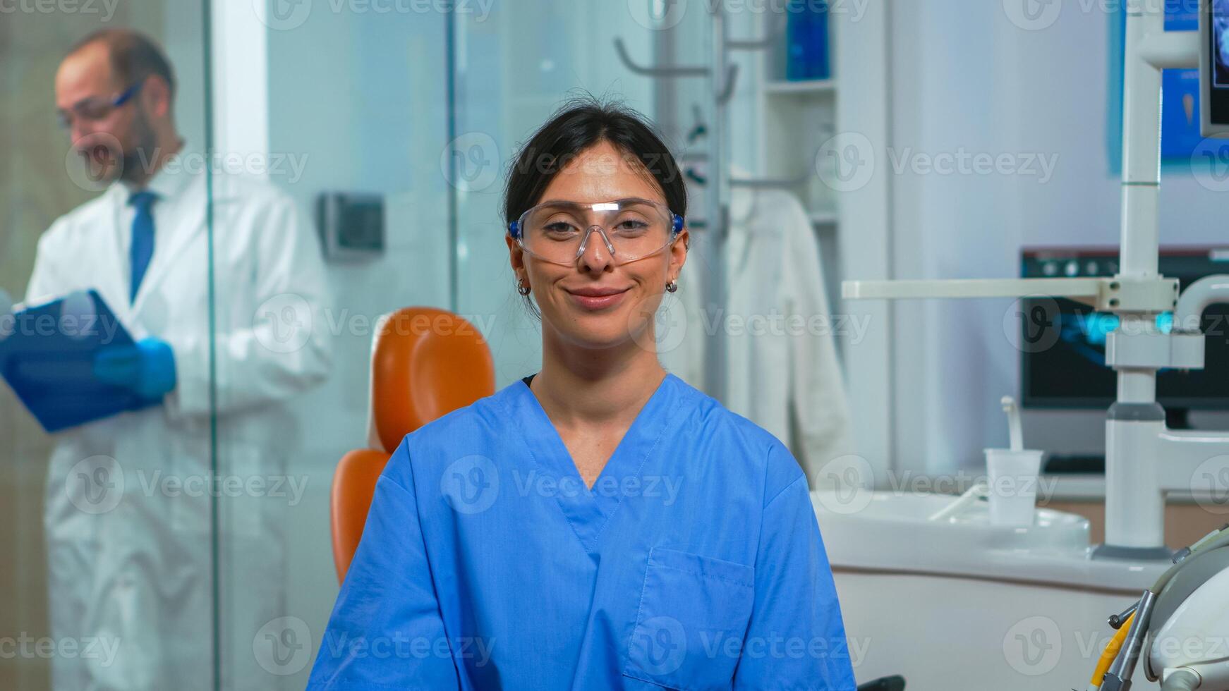 porträtt av leende sjuksköterska i dental kontor medan läkare är talande med patient i bakgrund. stomatolog assistent ser på webbkamera Sammanträde på stol i stomatologisk klinik. foto
