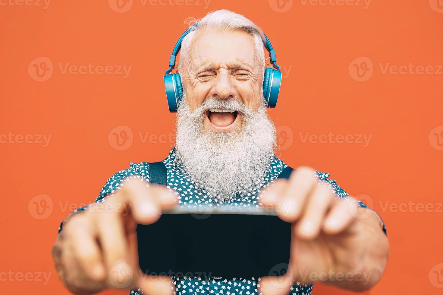 senior skäggig man framställning selfie med mobil telefon medan lyssnande till hans favorit Spellista med hörlurar - mode lagrad manlig har roligt med smartphone appar utomhus- - äldre, teknologi begrepp foto