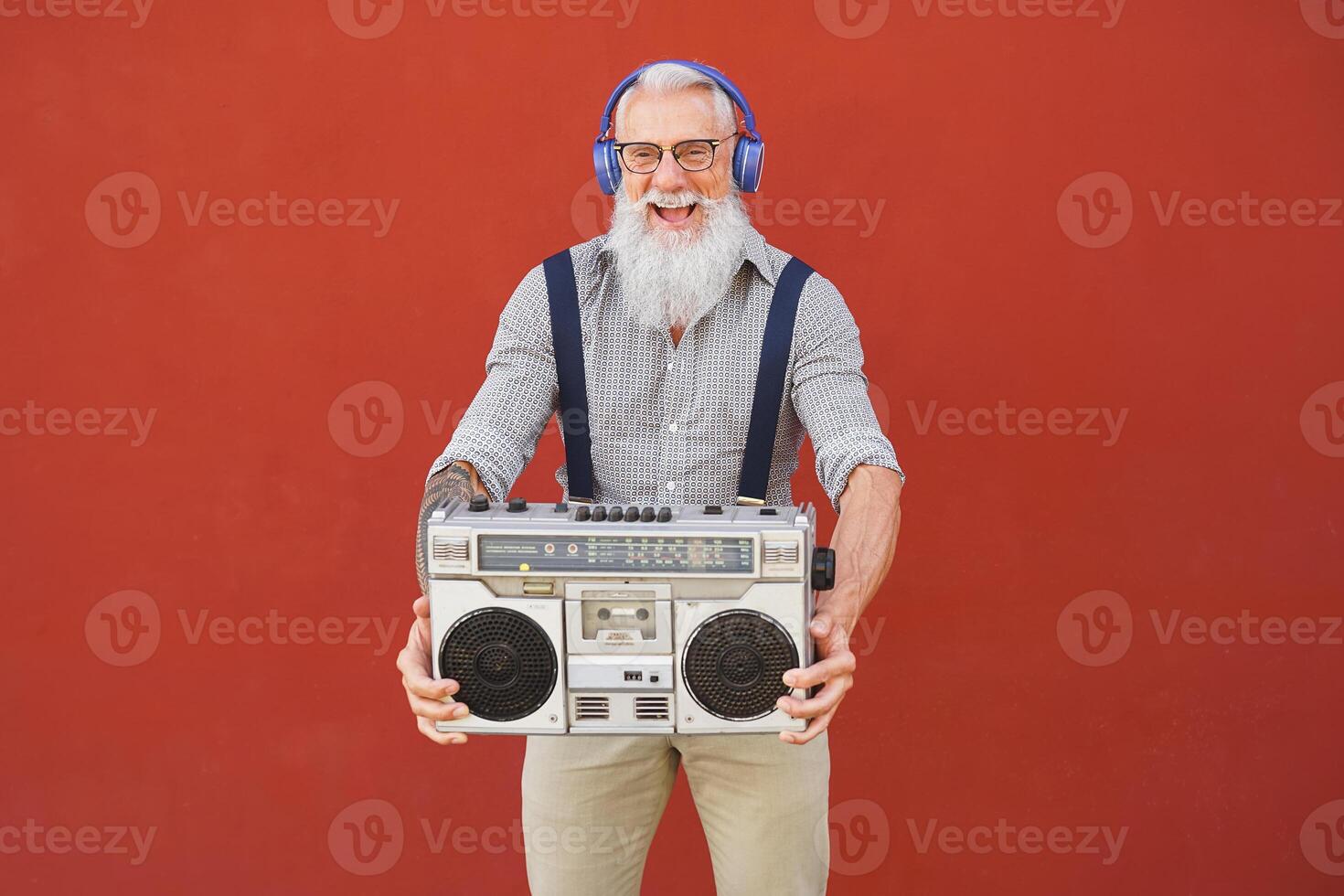 Lycklig senior man har roligt lyssnande till musik med trådlös hörlurar och årgång boombox utomhus- foto