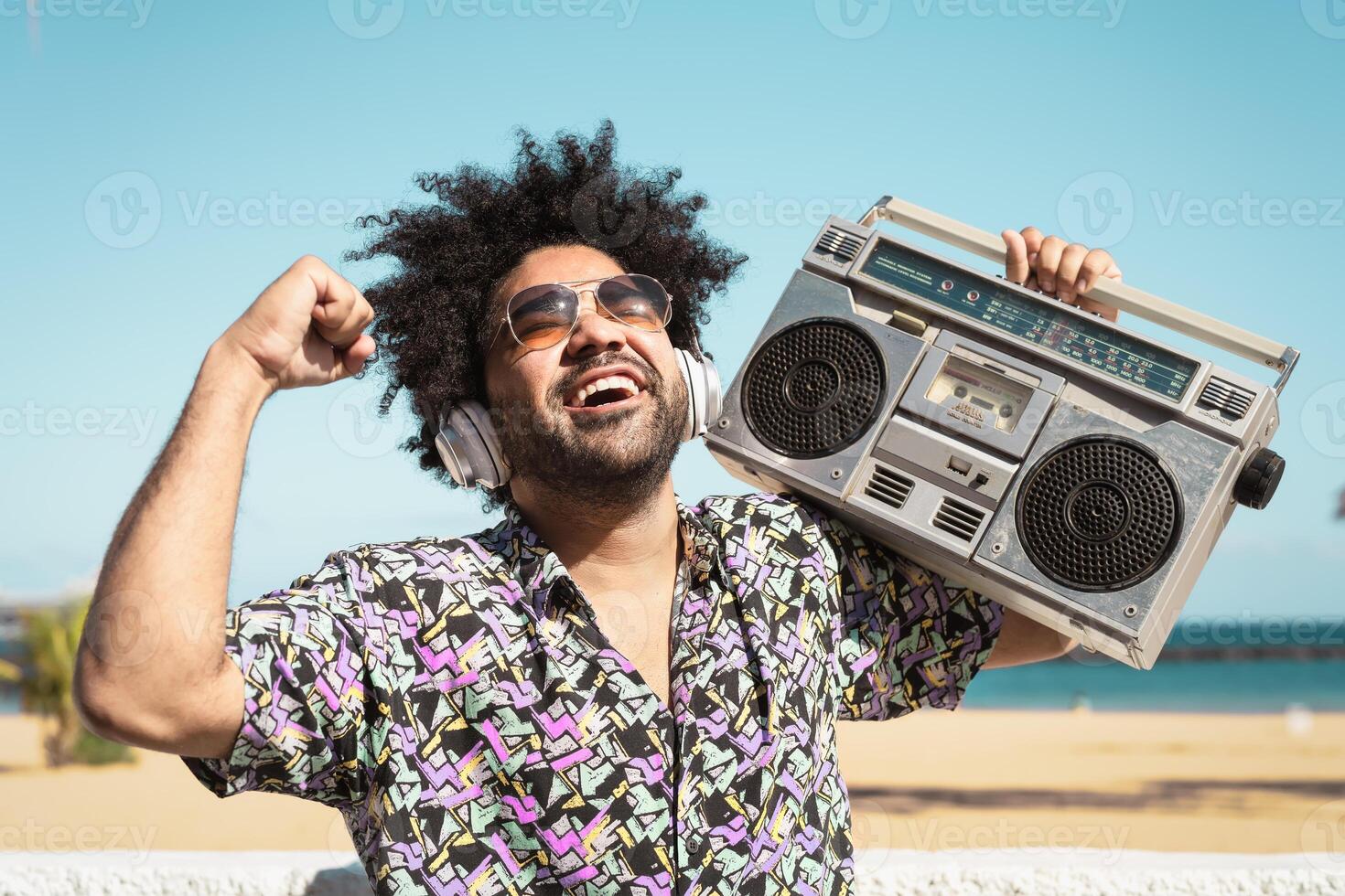 Lycklig afro man har roligt lyssnande till musik med trådlös hörlurar och årgång boombox på de strand under sommar tid foto