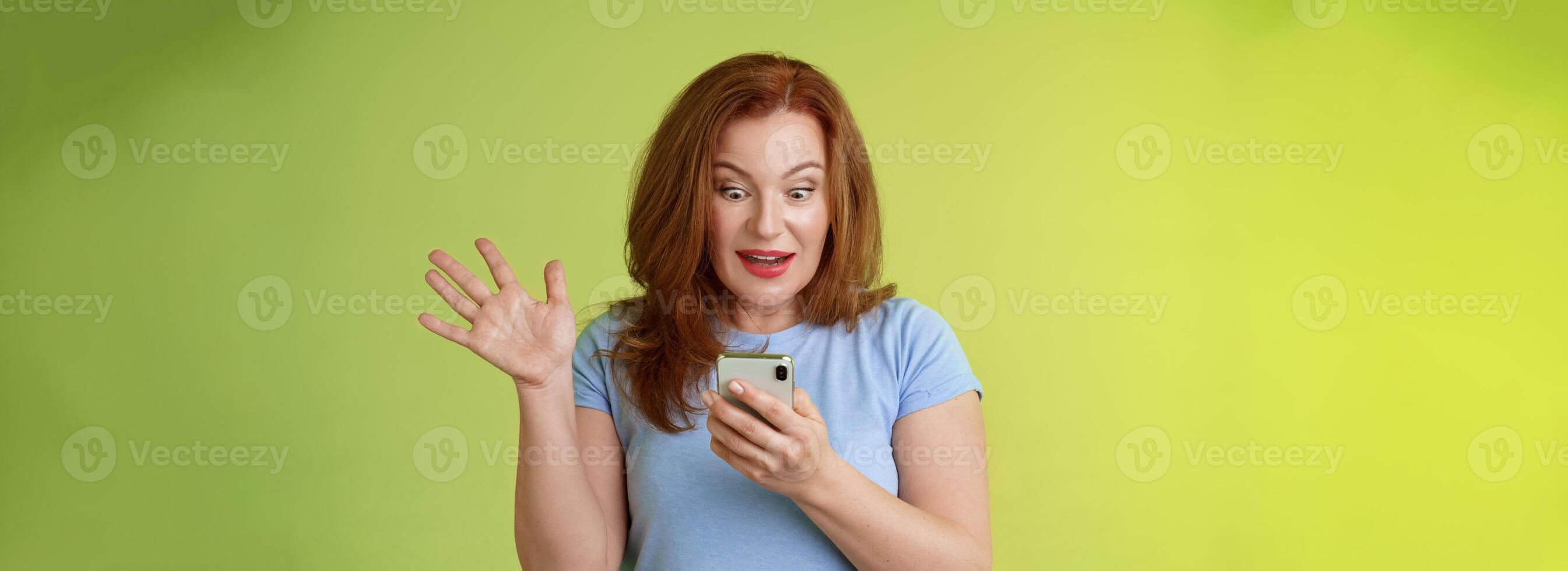 överraskad entusiastisk Lycklig rödhårig mogna medelålders kvinna motta excellent meddelande läsa perfekt Nyheter social media håll smartphone stirra förvånad nöjd mobil telefon skärm höja hand triumf foto
