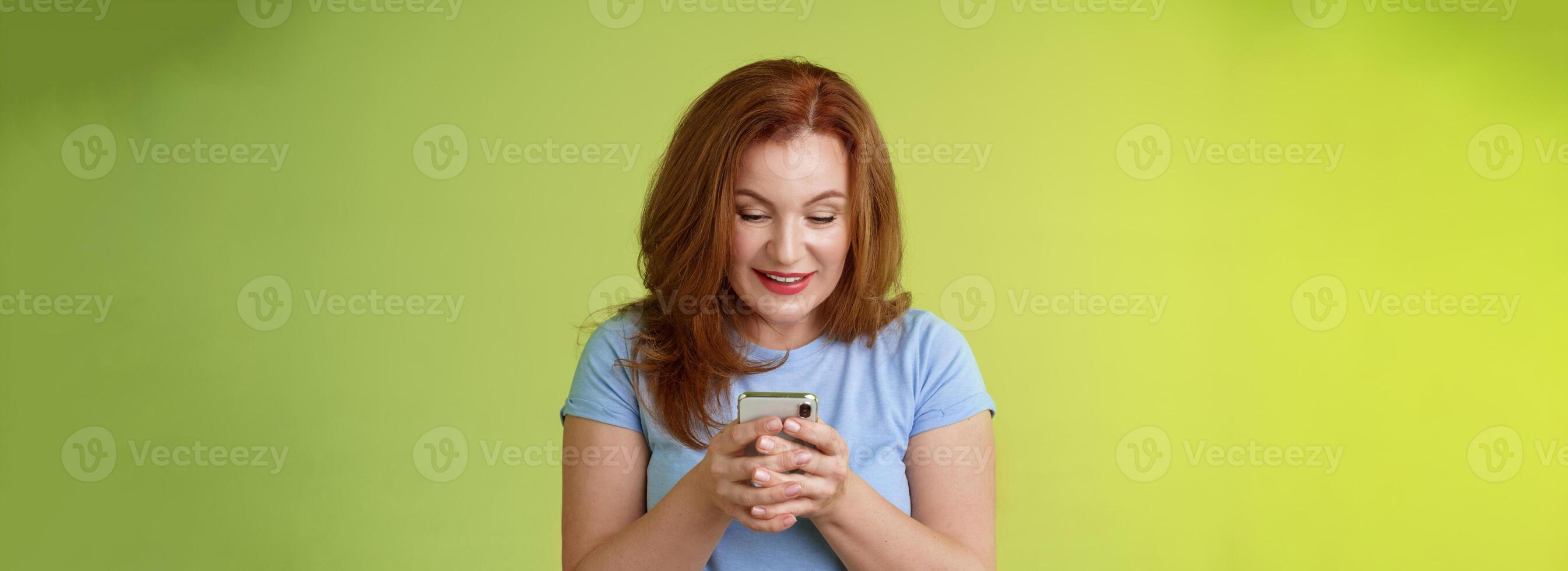 fascinerad upphetsad frestande rödhårig medelålders kvinna handla uppkopplad smartphone håll mobil telefon se nöjd underhöll grej skärm leende förtjust spelar spel hittades perfekt app foto