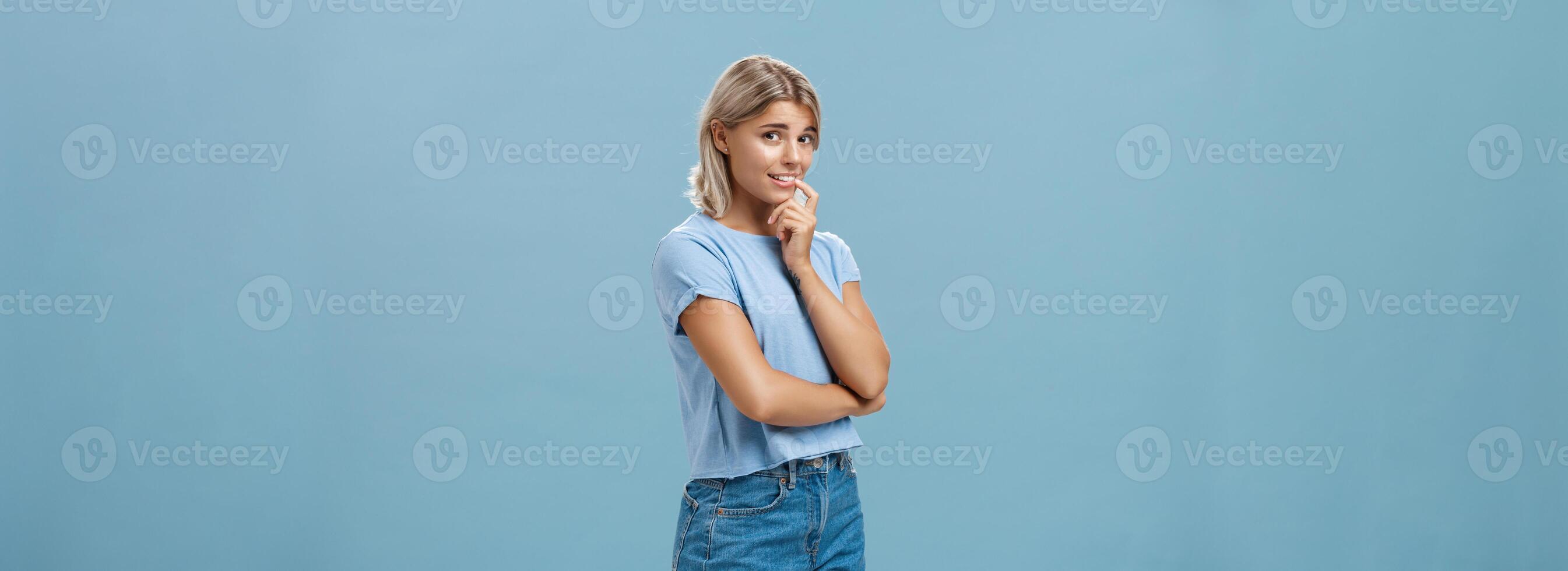 studio skott av osäker dumbom och osäker söt feminin flicka med blond hår stående halvvänt över blå bakgrund innehav finger på läpp medan varelse blyg fråga fråga leende tvekande foto