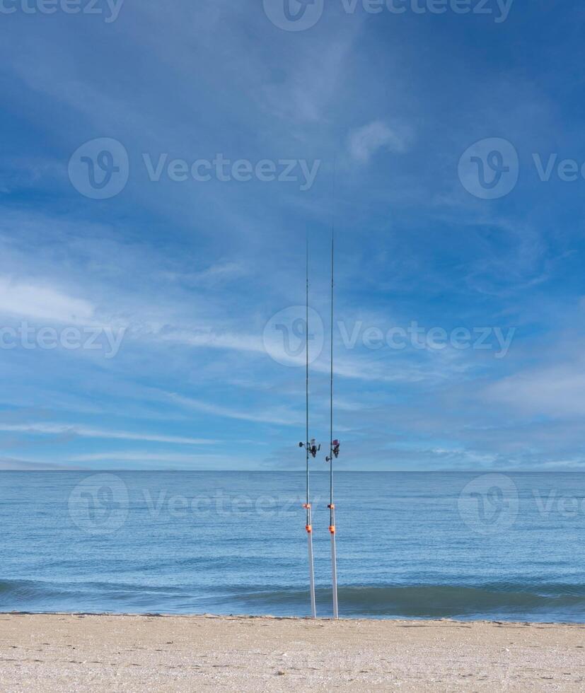 två fiske stavar, fastnat i de sand med de hav i de bakgrund och en molnig himmel foto