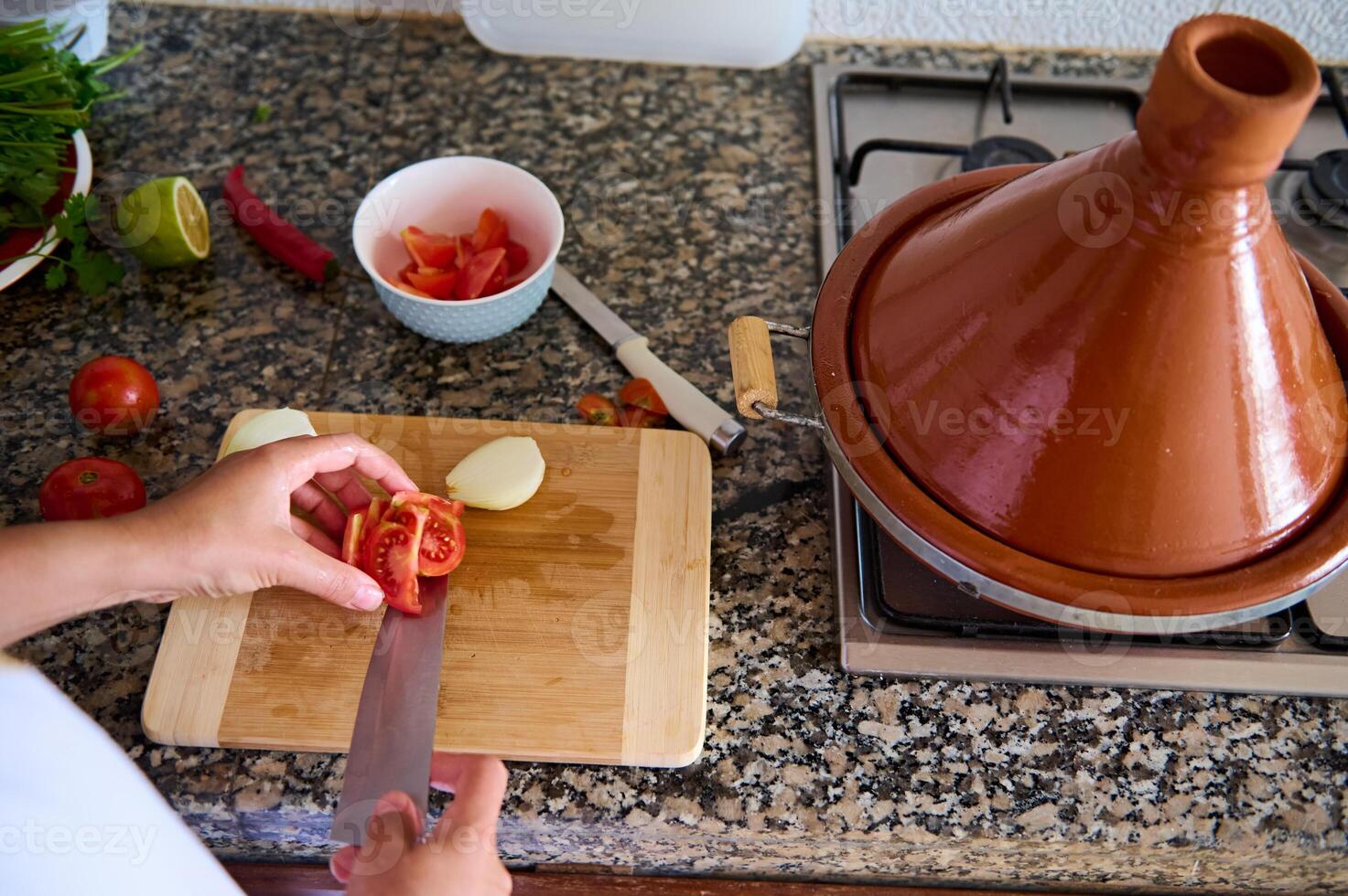 närbild se från ovan av en hemmafru hackning tomater på en skärande styrelse. en lera tajine pott på en stål spis foto