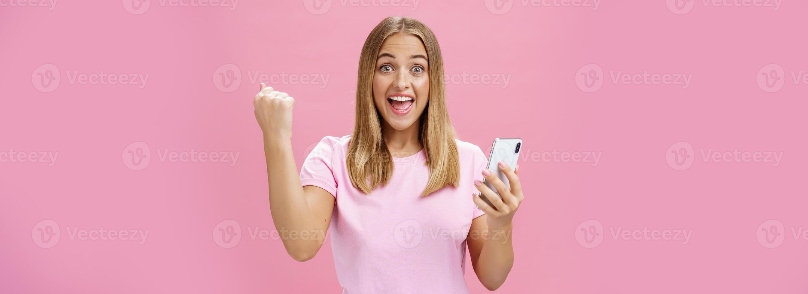 flicka slå egen spela in i smartphone spel höjning knöt näve i heja på och triumf innehav mobiltelefon, leende upphetsad och Lycklig på kamera fira seger med glad gest över rosa bakgrund foto