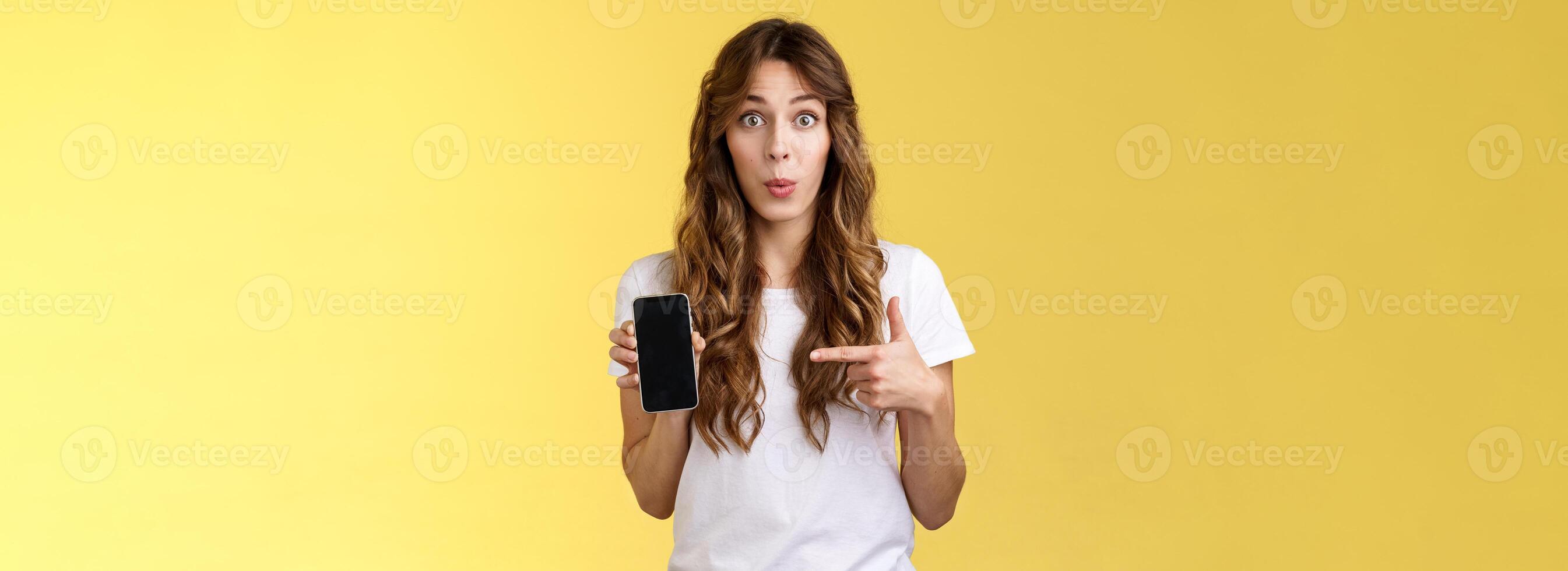 fängslande app kolla upp ut. entusiastisk överraskad attraktiv flickvän skvallrat vän ny pojkvän som visar nyfiken Foto smartphone håll mobil telefon pekande telefon skärm gul bakgrund