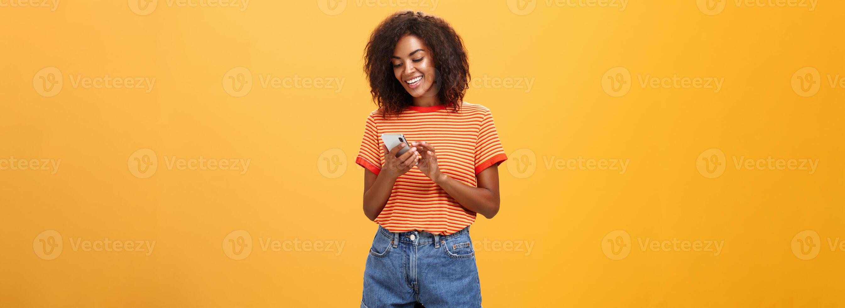 eleganta sorglös flicka textning vän komma över stående nöjd över orange vägg i eleganta denim shorts skriver meddelande eller rullning Nyheter i internet via smartphone gazing på enhet skärm med leende foto