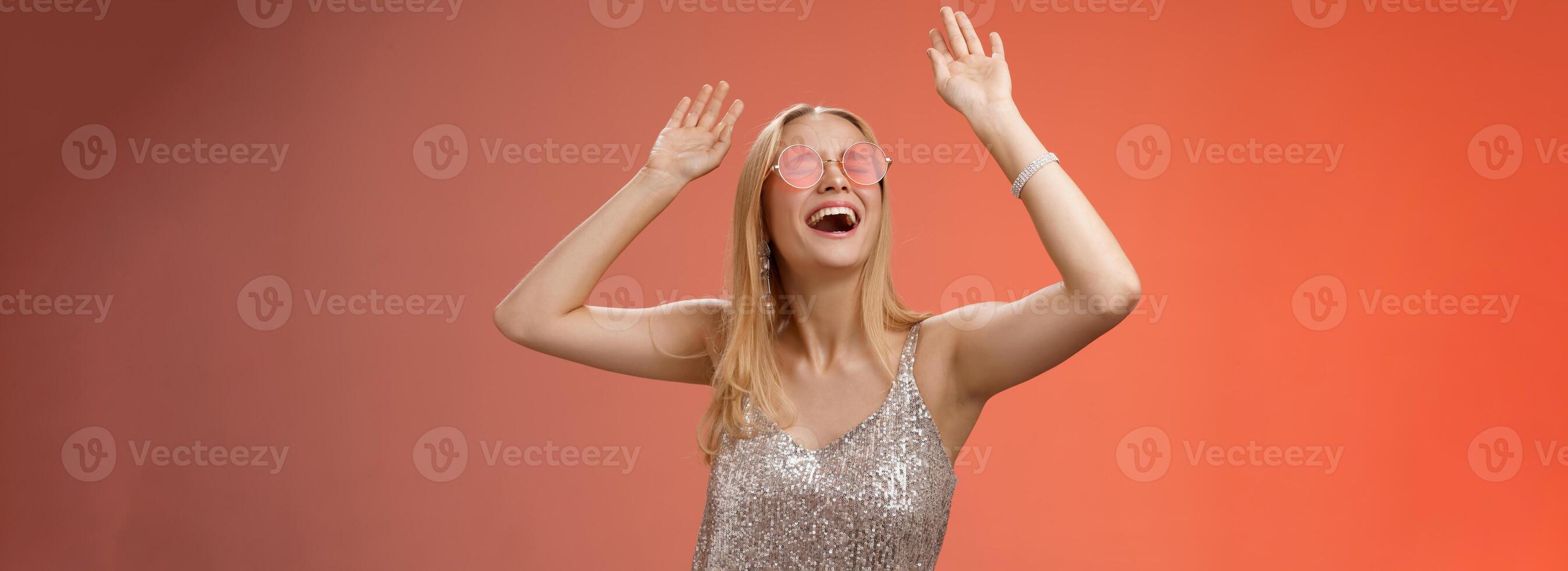 upphetsad kylning energiserad ung blond kvinna i silver- eleganta glittrande klänning solglasögon höja händer upp har roligt dans dansgolv nattklubb kasta fest fira födelsedag, röd bakgrund foto