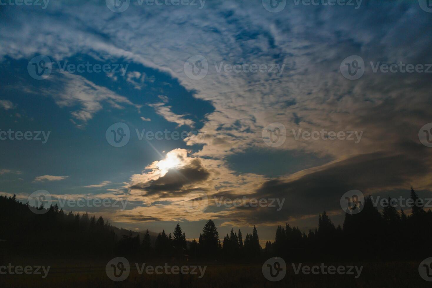 berg landskap tall träd nära dal och färgrik skog på sluttning under blå himmel med moln och dimma i måne ljus på natt foto