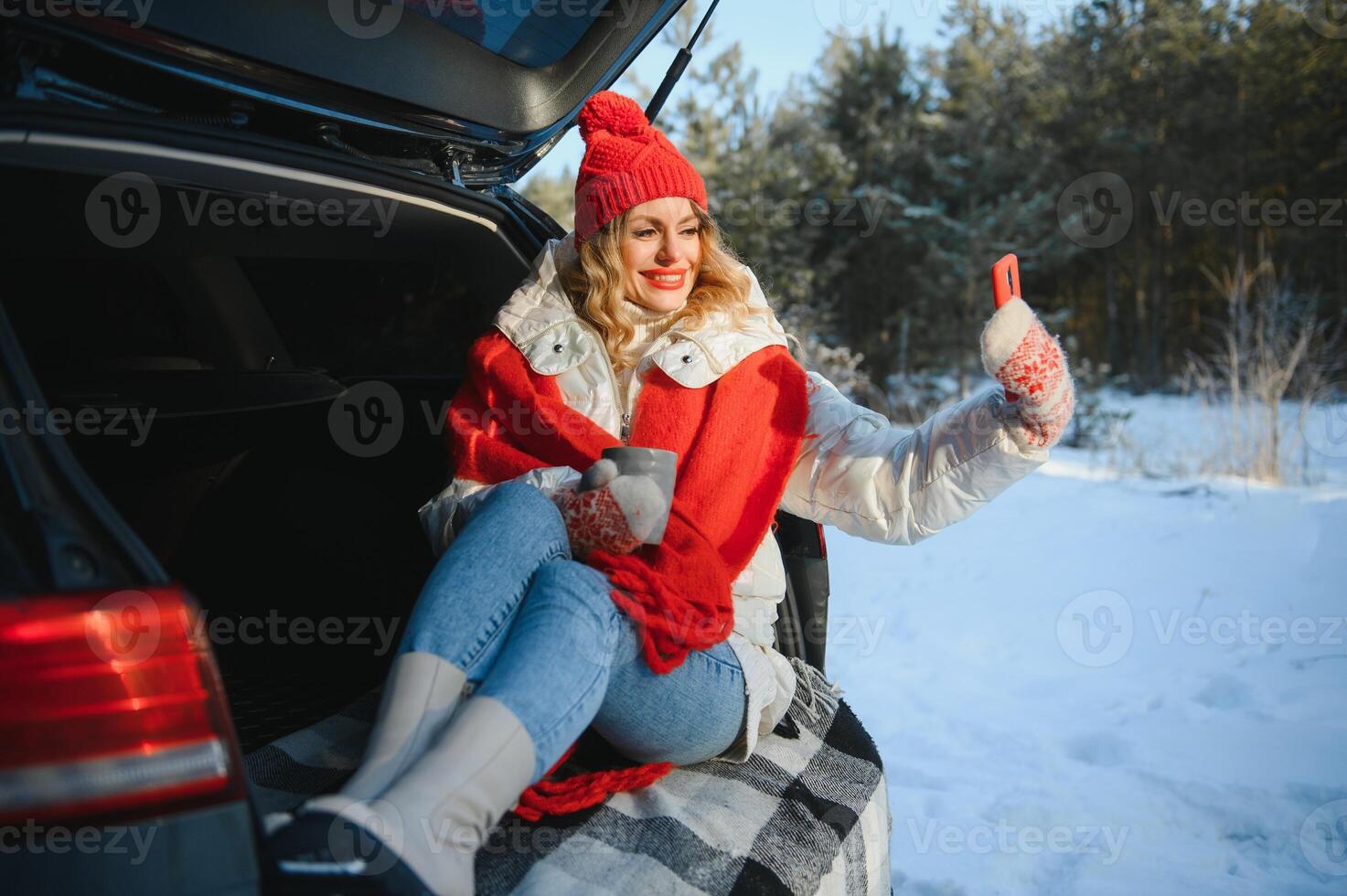 ung kvinna Sammanträde i bil trunk dricka värma te och tar en selfie på vinter- snöade dag. foto