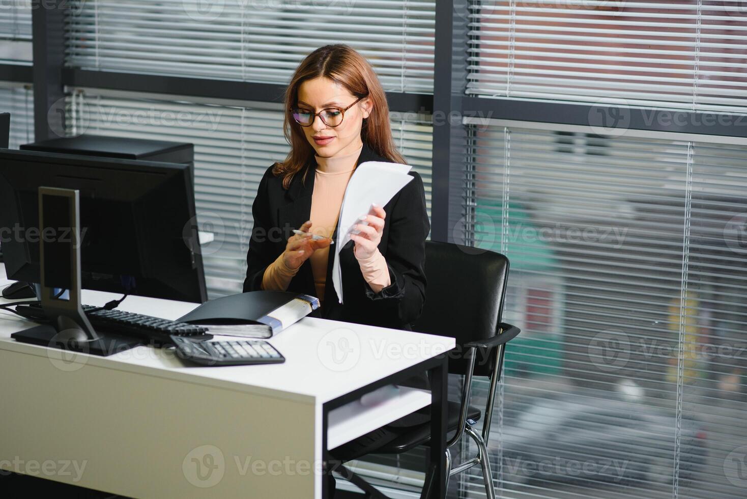 Söt, trevlig, söt, perfekt kvinna Sammanträde på henne skrivbord på läder stol i arbete station, bär glasögon, formell klädsel, har bärbar dator och anteckningsbok på de tabell foto
