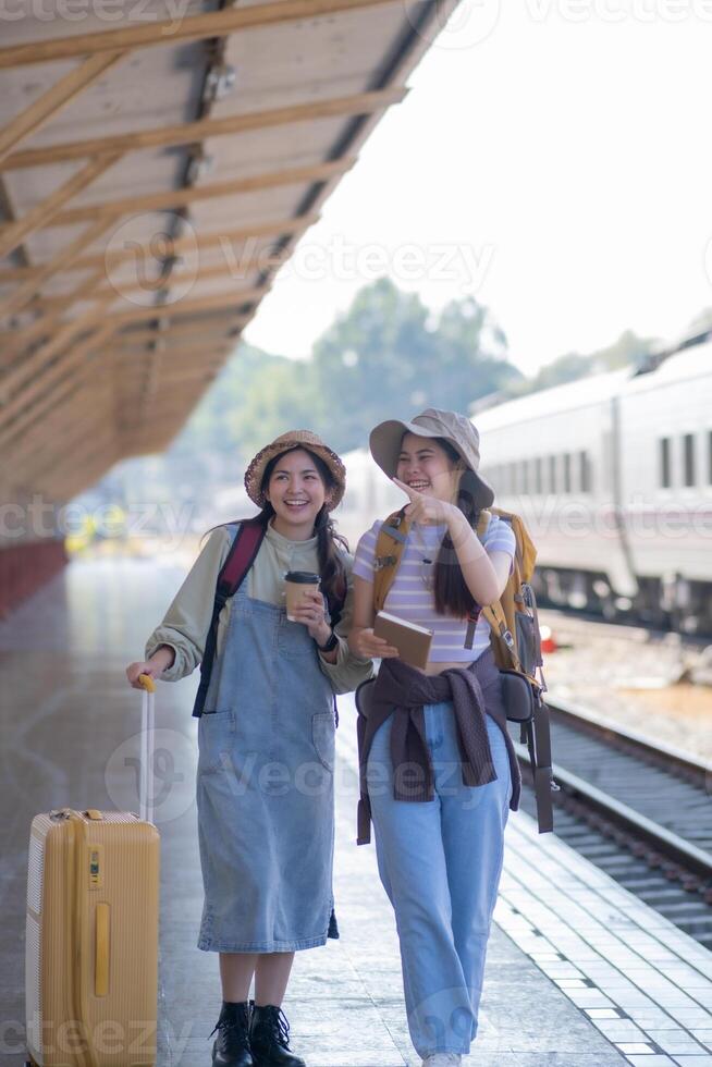 två ung asiatisk vänner flickor med ryggsäckar på järnväg station väntar för tåg, två skön kvinnor gående längs plattform på tåg station foto