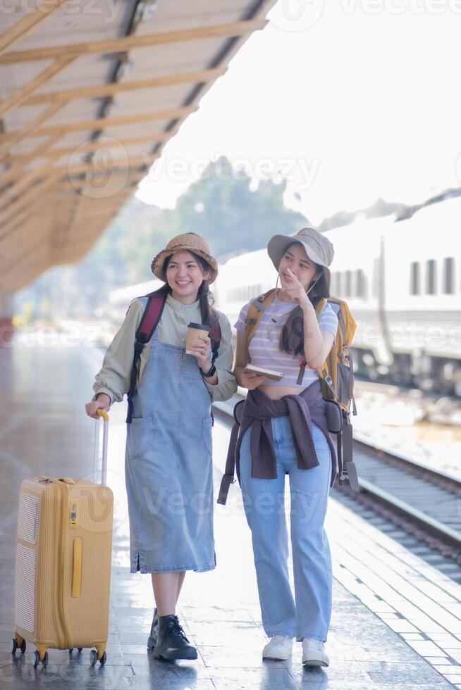 två ung asiatisk vänner flickor med ryggsäckar på järnväg station väntar för tåg, två skön kvinnor gående längs plattform på tåg station foto