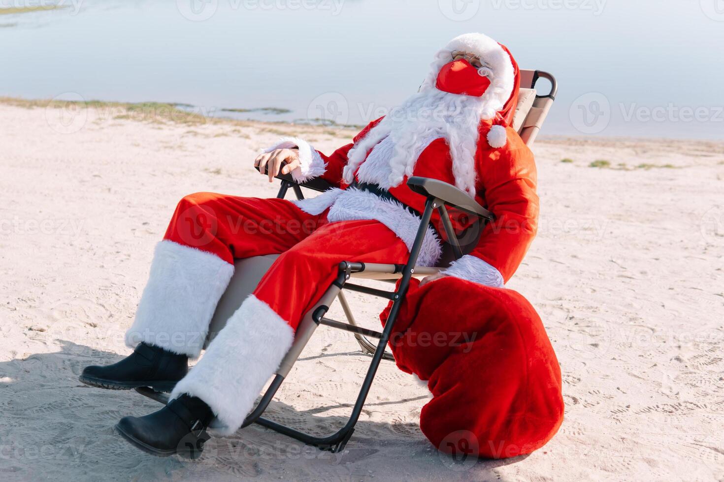 santa claus tar bort en röd medicinsk mask liggande på en Sol vilstol på de strand. slutet av de epidemi foto